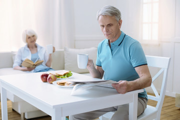 Concentrated man. Attentive senior man reading an interesting newspaper while his kind loving wife sitting by his side