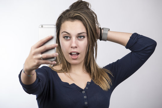 Bella ragazza bionda con vestito blu, si fa un selfie con espressione molto sicura con la bocca aperta - sfondo chiaro