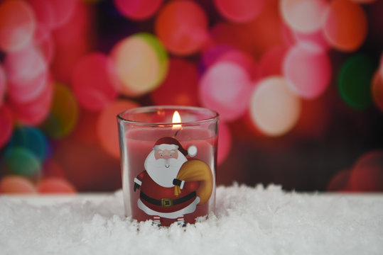 Christmas photography picture of red glass candle with lit flame and happy jolly Santa Claus design and laid in snow with warm cozy red pink christmas light background