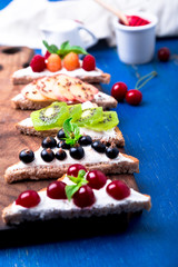 Fruit toast on wooden board on blue rustic background. Healthy breakfast. Clean eating. Dieting concept. Grain bread slices with cream cheese and various fruit, berries, seeds. Vegetarian. Top view.