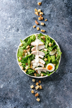 Classic Caesar salad with grilled chicken breast and half of egg in white ceramic plate. Served with croutons and dressing over dark gray texture background. Top view, space.
