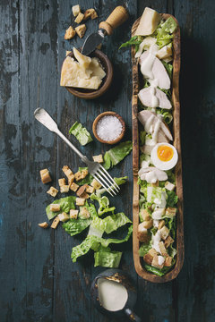 Classic Caesar salad with grilled chicken breast and half of egg in olive wood bowl. Served with ingredients above over old dark blue wooden background. Top view, space. Rustic style