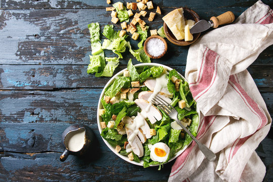 Classic Caesar salad with grilled chicken breast and half of egg in white ceramic plate. Served on towel with ingredients above over old dark blue wooden background. Top view, space. Rustic style