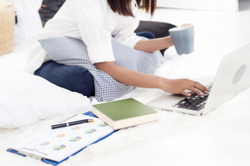 The lady in blurry light design background of the lady in white shirt and blue jean is workng at home by use laptop beside paper business investment chart and hold coffee cup in left hand,ading book