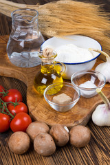 Frische Zutaten für einen traditionellen Pizzateig und Belag mit Tomaten, Pilzen und Knoblauch auf einem Brett aus Olivenholz mit Getreideähren im Hintergrund