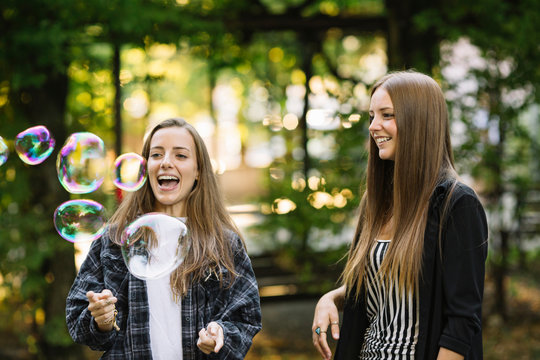 Two young female friends bursting floating bubbles in park