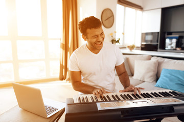 A black man sits in the living room of his apartment and plays a synthesizer. He composes music.