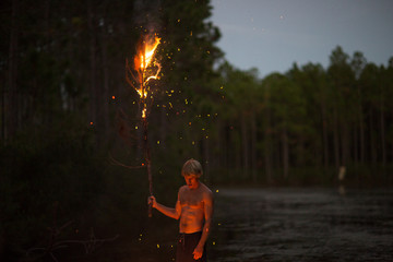 Young man standing near water, at dusk, holding burning tree branch