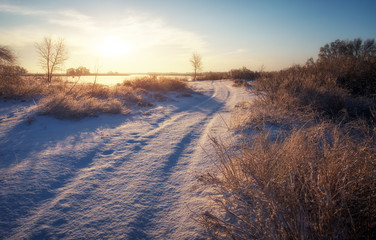 Winter snow-covered road, plants and trees in the frost, coast of river and sun at sunset.