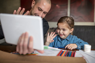 Man and daughter at table waving at digital tablet