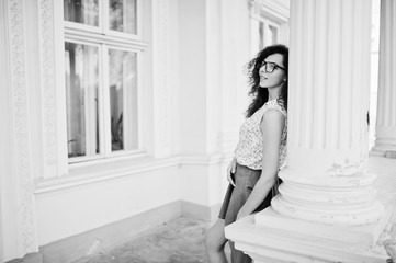 Curly stylish girl wear on blue jeans skirt, blouse and glasses posed near old vintage house.