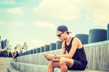 Fototapeta na wymiar Man travels in New York in hot summer. Wearing black tank top, shorts, cap worn backward, young guy sits on wooden stairs at park, reads messages on cell phone. Filtered effect..