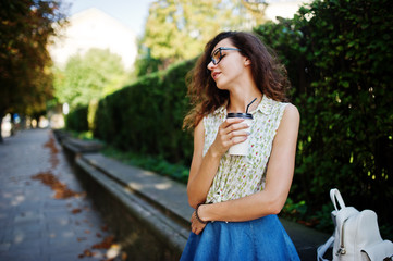 Fototapeta na wymiar Curly stylish girl wear on blue jeans skirt, blouse and glasses posed at street of city with cup of coffee.