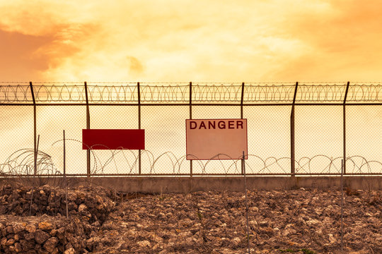 metal mesh wall with barbed wires  and rolled razor barbed wires security fence and danger notice board on sunset background