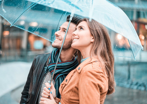 Young happy couple under the rain day covering with transparent umbrella in city center - Lovers traveling Europe during fall season - Love concept - Focus on woman face - Teal and orange filter