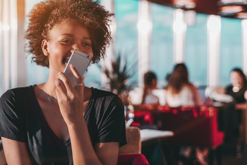Appealing young Brazilian female is sitting in restaurant and laughing while dictating audio...