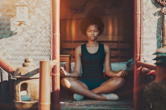 Young charming African American female with curly hair sitting inside of oriental pagoda on a matting and meditating, halo appeared around her head, she is feeling peaceful and calm, reached nirvana