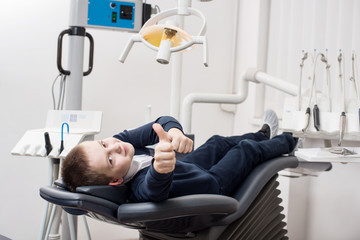 Happy patient boy showing thumbs up at dental office. Medicine, stomatology and health care concept. Dental equipment