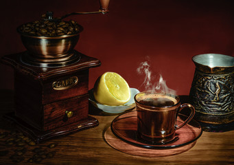 A Cup of hot coffee, steam over a Cup, lemon, boiling water, manual coffee grinder, coffee beans on the table. The dark maroon background.