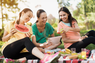 Three pregnant women rest on nature after doing yoga. One of them holds a tablet