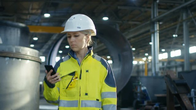Female Industrial Worker in the Hard Hat Uses Mobile Phone While Walking Through Heavy Industry Manufacturing Factory. In the Background Various Metalwork Project Parts Lying 