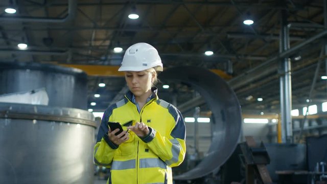 Female Industrial Worker in the Hard Hat Uses Mobile Phone While Walking Through Heavy Industry Manufacturing Factory. In the Background Various Metalwork Project Parts Lying 