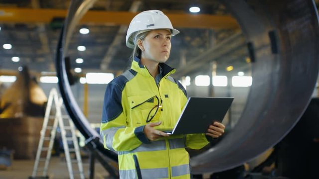 Female Industrial Engineer in the Hard Hat and Safety Jacket Uses Laptop Computer while Standing in the Heavy Industry Manufacturing Factory. In the Background Various Metalwork Project Parts Lying 