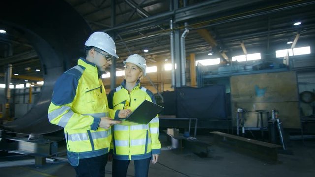 Male and Female Industrial Engineers in Hard Hats Discuss New Project while Using Laptop. They Work in the  Heavy Industry Manufacturing Factory with Metalwork Components Lying Around. Arc View Shot.