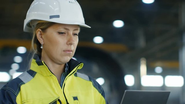 Portrait Female Industrial Engineer in the Hard Hat Uses Laptop Computer while Standing in the Heavy Industry Manufacturing Factory. In the Background Various Metalwork Project Parts Lying 