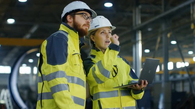 Male and Female Industrial Engineers in Hard Hats Discuss New Project while Using Laptop. They Make Showing Gestures.They Work at the Heavy Industry Manufacturing Factory. Long Shot.