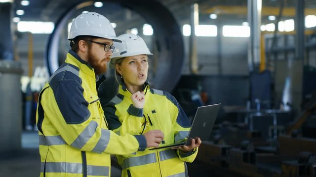 Male and Female Industrial Engineers in Hard Hats Discuss New Project while Using Laptop. They Make Showing Gestures.They Work in a Heavy Industry Manufacturing Factory. Long Shot.