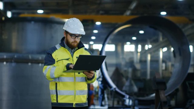 Industrial Engineer in Hard Hat Wearing Safety Jacket Uses Laptop. He Works in the Heavy Industry Manufacturing Factory with Various Metalworking Processes are in Progress.