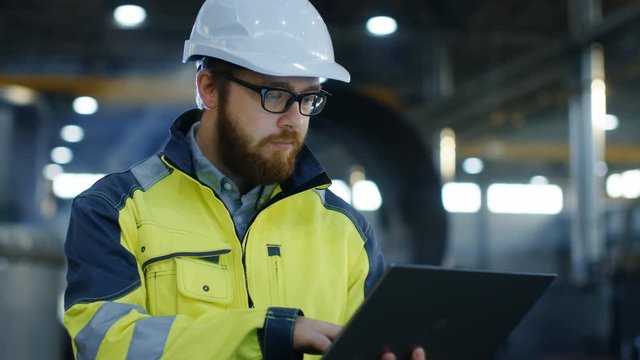 Industrial Engineer in Hard Hat Wearing Safety Jacket Uses Laptop. He Works in the Heavy Industry Manufacturing Factory with Various Metalworking Processes are in Progress.