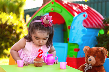 Baby toddler girl in outdoor second birthday party blowing candle on muffin. Teddy Bear as best...