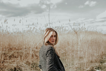 Pretty and young woman with long blonde hair dressed in wool coat looking at camera over her shoulder and smiling. Background bulrush. Autumn. Outdoor. Medium shot