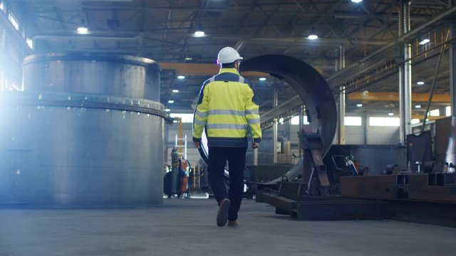 Industrial Engineer in Hard Hat Wearing Safety Jacket Walks Through Heavy Industry Manufacturing Factory with Various Metalworking Processes.