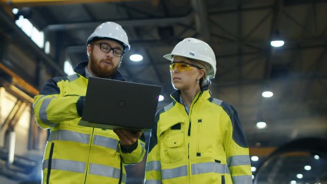 Male and Female Industrial Engineers use Laptop and Have Discussion While Walking Through Heavy Industry Manufacturing Factory. They Wear Hard Hats and Safety Jackets. 