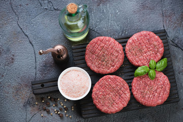 Black wooden chopping board with raw fresh beef burger cutlets and condiments on a cracked asphalt background, above view