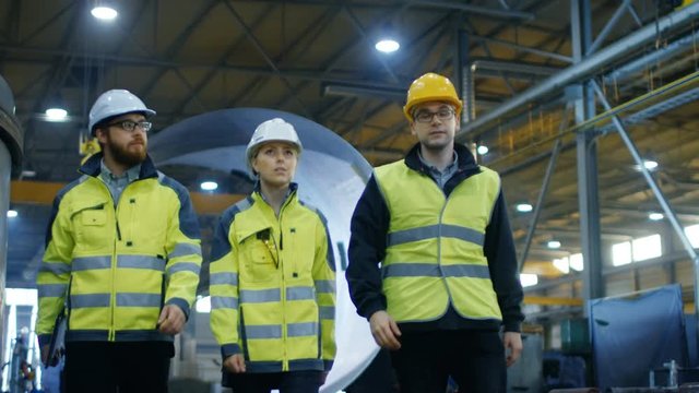 Following Shot of Three Engineers Walking Through Heavy Industry Manufacturing Factory. In the Background Welding Work in Progress, Various Metalwork, Pipeline/ Barrel Components. Slow Motion.