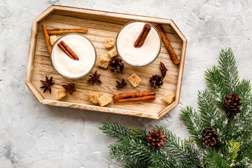 Homemade classic eggnog with cinamon and badian on wooden tray near spruce branch, pinecones on grey stone background top view