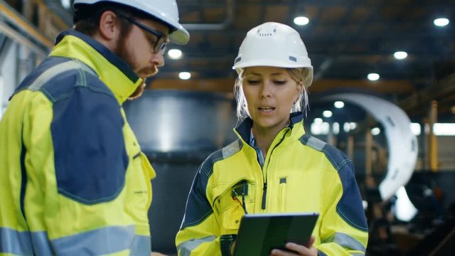 Male and Female Industrial Engineers in Hard Hats and Safety Jackets Discuss New Project while Using Tablet Computer. They Work at the Heavy Industry Manufacturing Factory. 