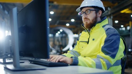 Industrial Engineer Works at Workspace on a Personal Computer.  He Wears Hard Hat and Safety Jacket and Works in the Main Workshop of the Heavy Industry Manufacturing Factory. - Powered by Adobe