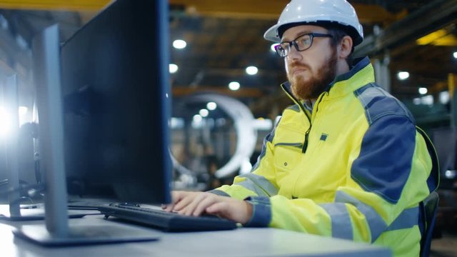 Industrial Engineer Works at Workspace on a Personal Computer.  He Wears Hard Hat and Safety Jacket and Works in the Main Workshop of the Heavy Industry Manufacturing Factory.