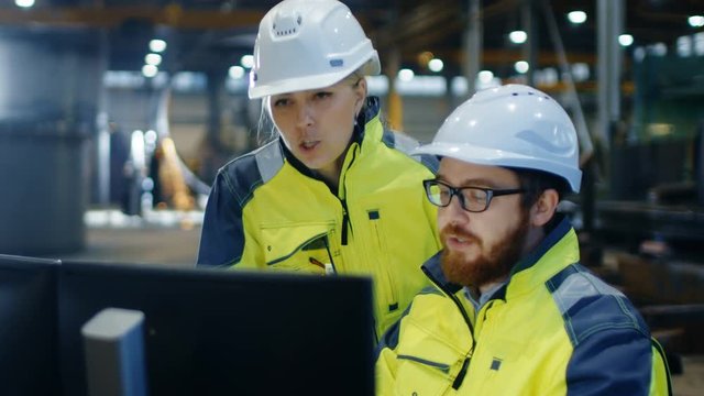 Male Industrial Engineer Works on the Personal Computer while Female Factory Worker Talks about Project Details. They Work in Heavy Industry Manufacturing Facility