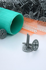 A set of construction items for the insulation of walls. Plastic dowels, a roll of mesh for the insulation of facades and a corner profile with a grid lie on the foam polystyrene surface