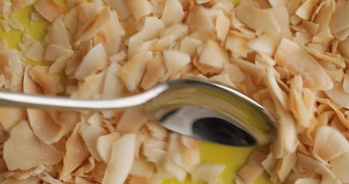 Close video of using a spoon to mix and get a spoonful of freshly toasted coconut flakes from the bottom of a bright yellow bowl.