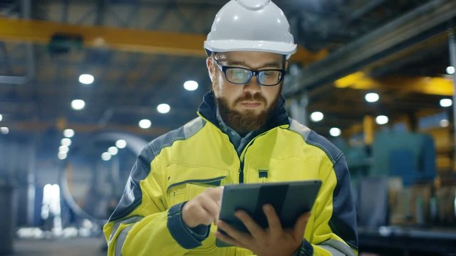 Industrial Engineer in Hard Hat Wearing Safety Jacket Uses Touchscreen Tablet Computer. He Works at the Heavy Industry Manufacturing Factory.