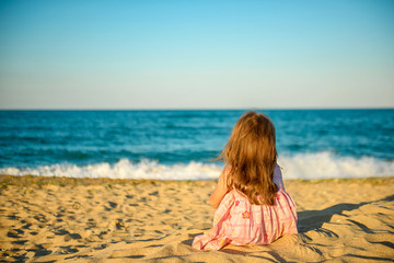 little girl in a dress sitting on the beach and dreamily looking at the sea