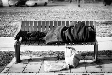 Homeless refugee man in black coat sleeps on the bench on the street black and white