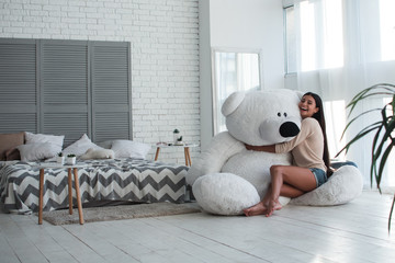 Perfect present. Beautiful young woman hugging huge teddy bear and looking at camera with smile while sitting indoors - obrazy, fototapety, plakaty
